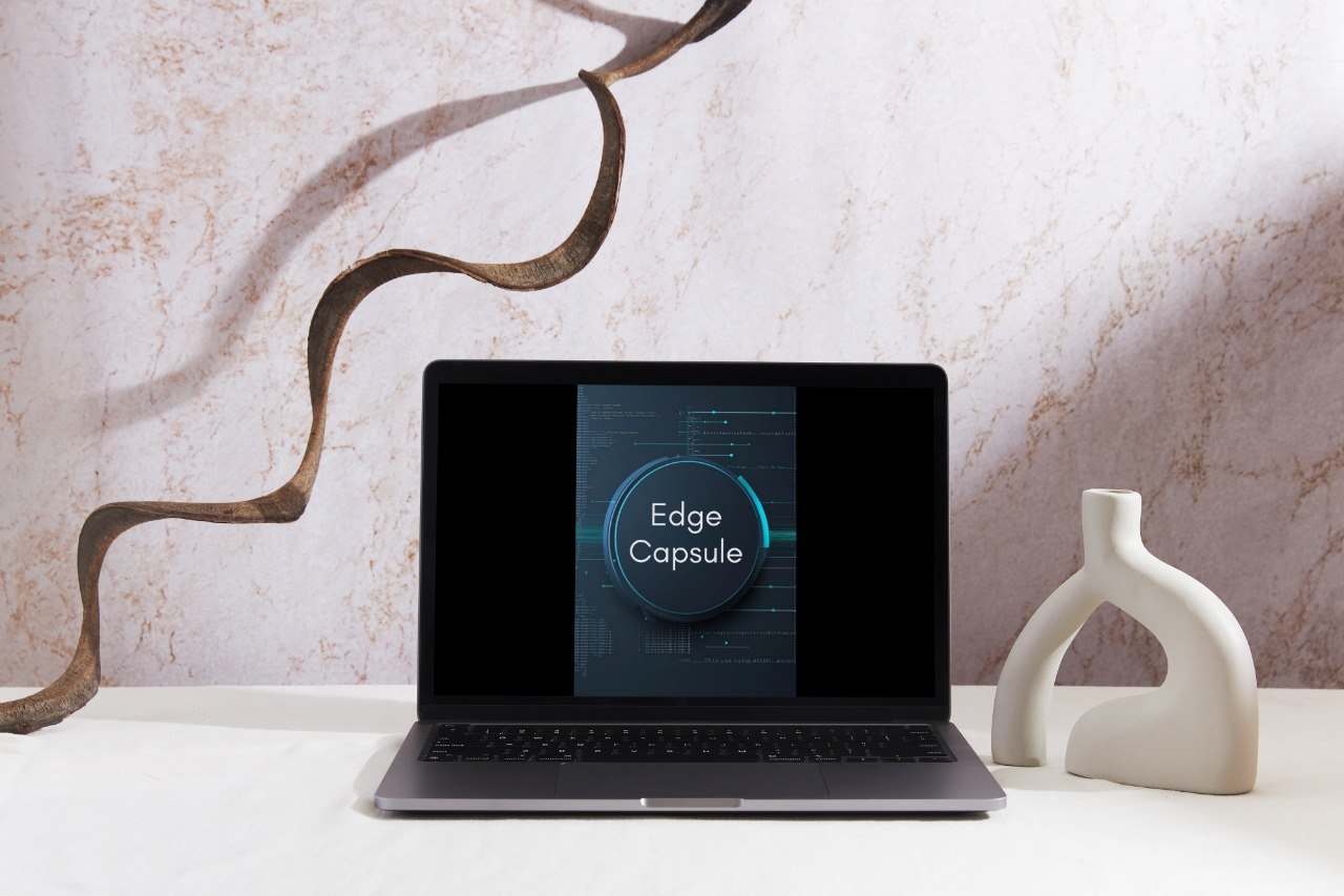 A laptop sits open on a white surface against a textured light wall, displaying a dark interface labeled 'Edge Capsule.' Decorative objects flank the laptop: a sculptural brown branch-like form on the left and a white abstract vase on the right.