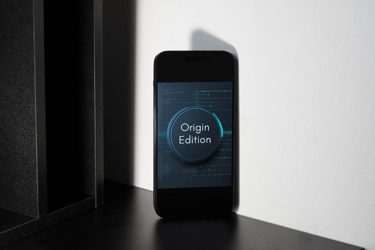 A black smartphone standing upright on a dark surface against a white wall, displaying a blue and black screen with the text 'Origin Edition' in a circular graphic.