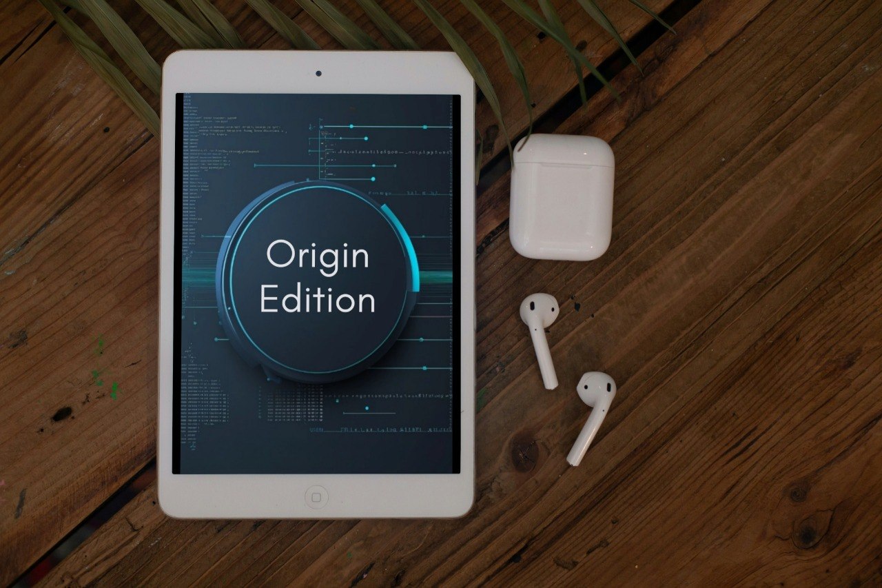 A white tablet displaying a dark blue 'Origin Edition' graphic, arranged on a wooden surface next to white wireless earbuds and their charging case.
