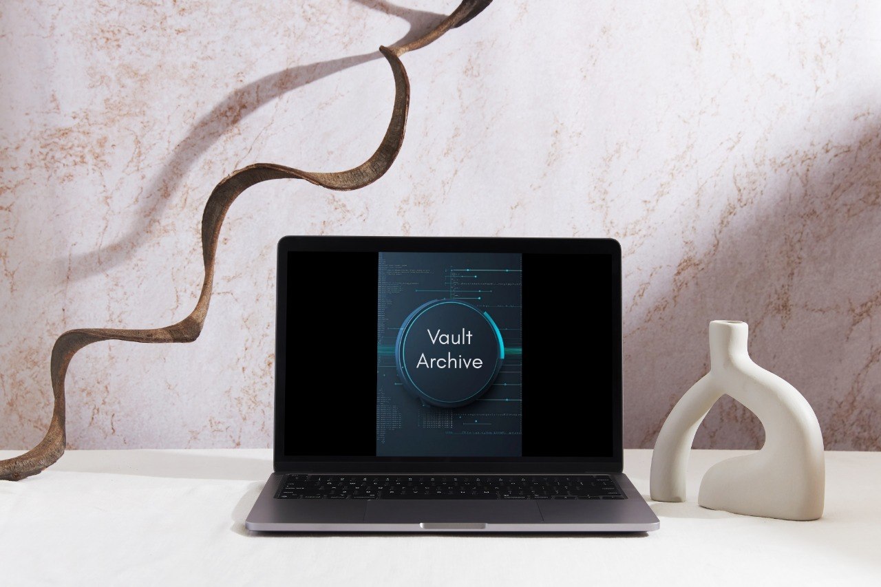 A laptop sits open on a white surface against a textured beige wall, displaying a screen with the words 'Vault Archive' inside a circular design. Decorative objects, including a curved brown metal sculpture and a white ceramic vase, frame the laptop.